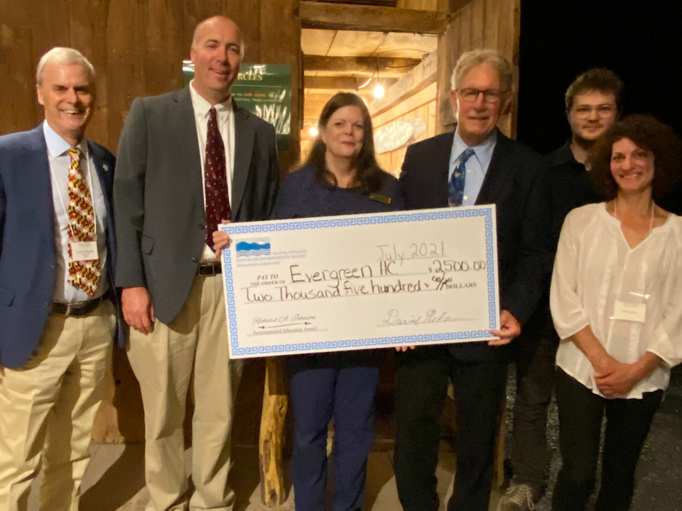 From left to right: Dr. Peter Goodwin, Dr. David Nelson, Janice Keene holding large prop check, Francis "Champ" Zumbrun, Adron Fiscus, and Marta Fiscus. All are standing in barn, facing camera. 