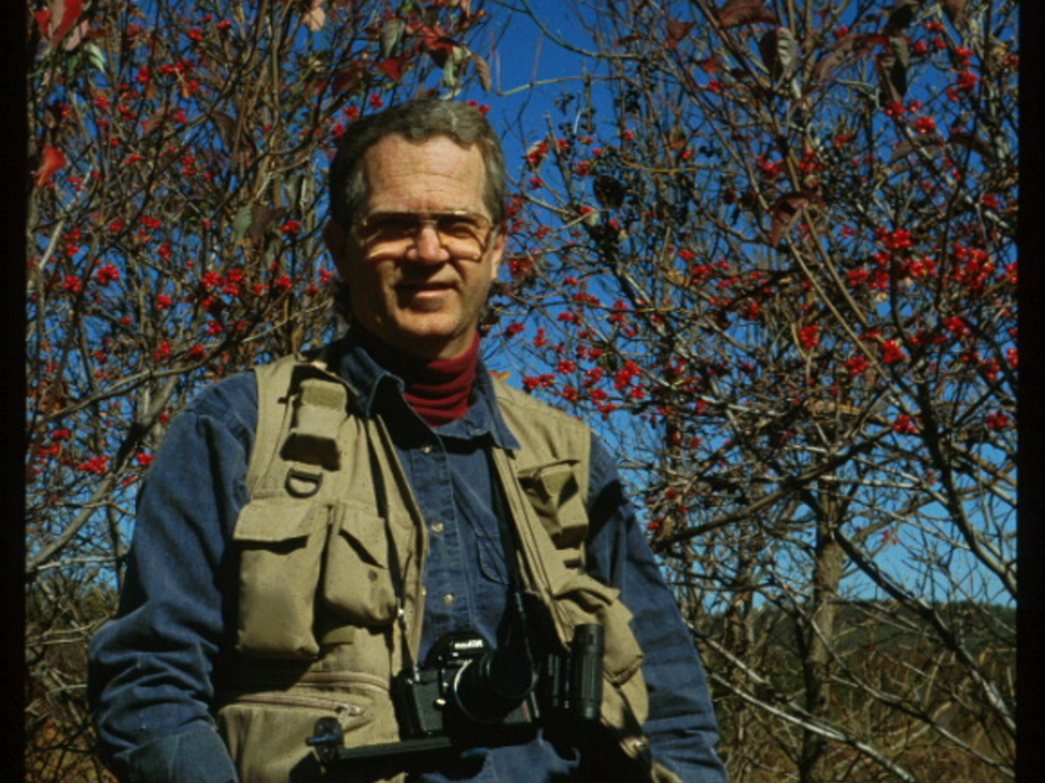 Dr. Richard A. Johnson outdoors surrounded by trees with binoculars and other birding equipment,