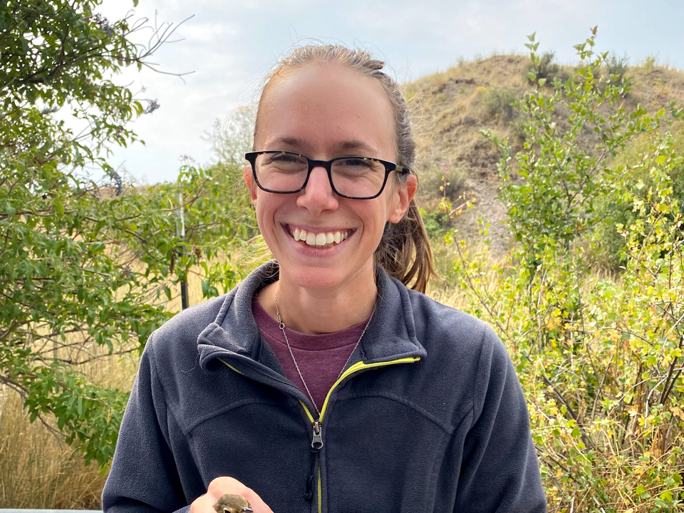 Dr. Joely DeSimone in blue fleece zippered jacket holding bird with trees and plants in background. 