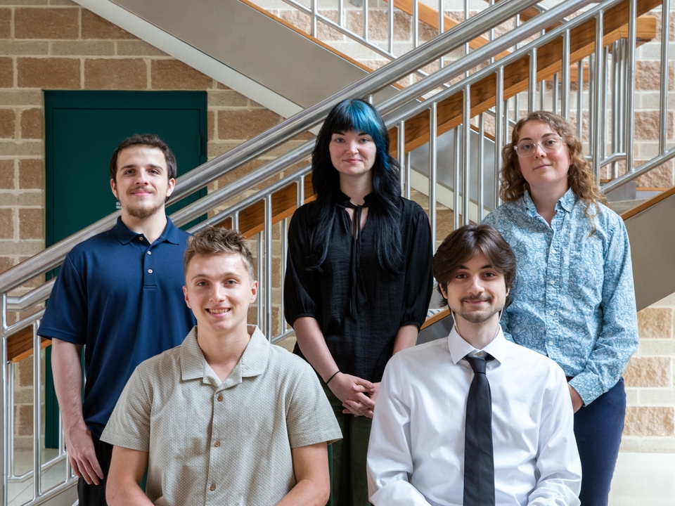 Two male interns seated with one male and two female interns standing behind them. Metal stairway and brick wall in the background. 