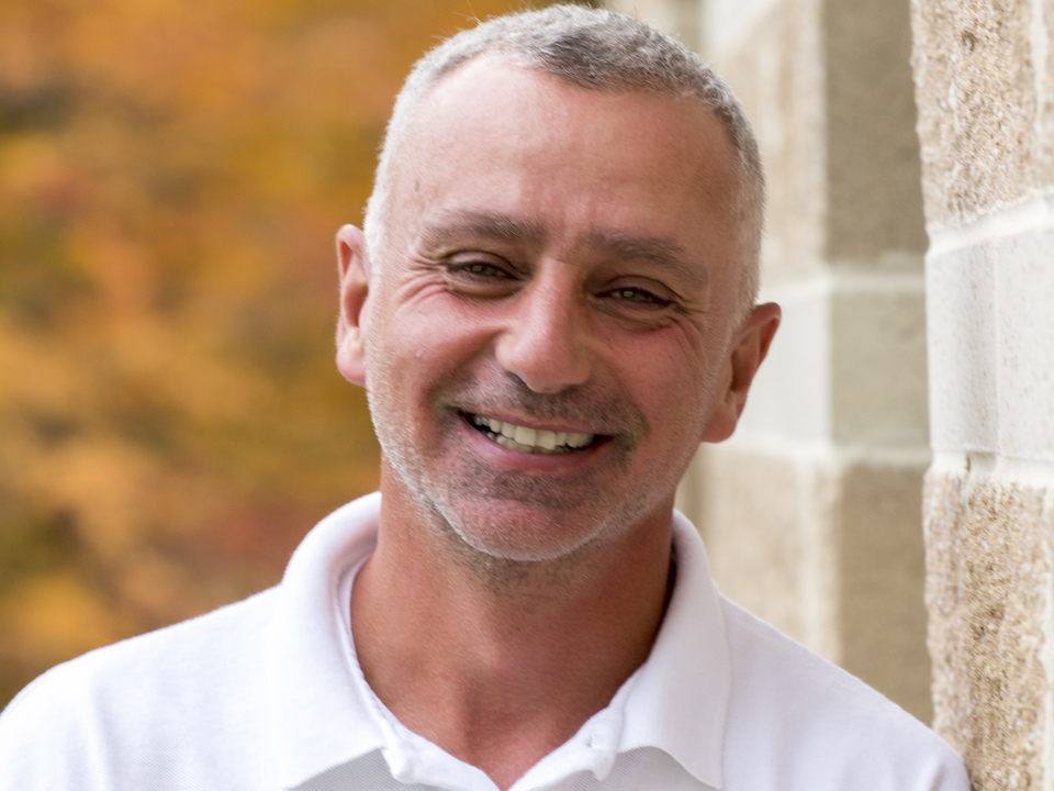 Headshot of John Piasecki in white shirt leaning against brick building with orange fall leaves in background. 