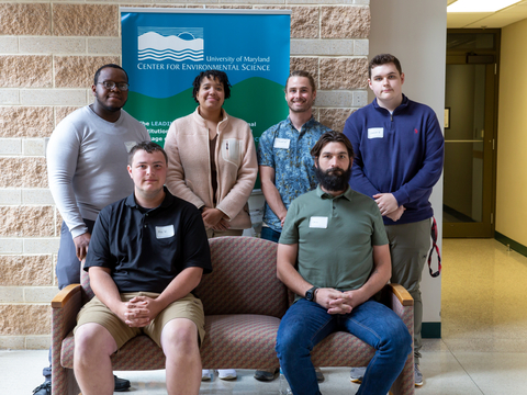 Six Appalachian Laboratory interns. Four standing behind two seated on pink sofa. Brick colored stone wall in background with UMCES logo. 