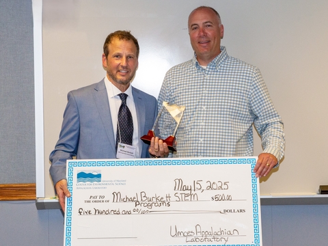 Mick Burkett and David Nelson holding inspiration award and ceremonial check with white marker board in background. 