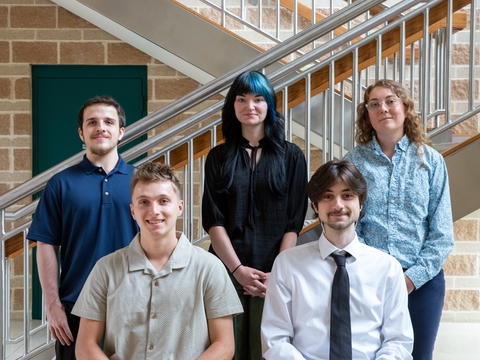 Two male interns seated with one male and two female interns standing behind them. Metal stairway and brick wall in the background. 