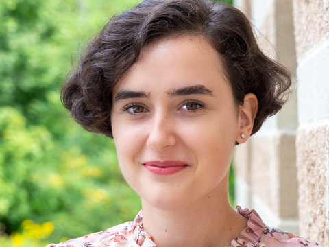 Haedshot of Mihaela Borota in pink floral blouse leaning against stone wall with blurred greenery in background. 