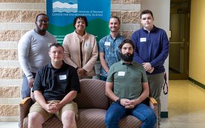Six Appalachian Laboratory interns. Four standing behind two seated on pink sofa. Brick colored stone wall in background with UMCES logo. 
