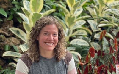 Annie Stark wearing a grey sweater with pink, white and grey striped sleeves with large green plants in background. 