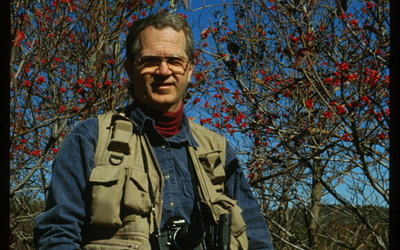 Dr. Richard A. Johnson outdoors surrounded by trees with binoculars and other birding equipment,