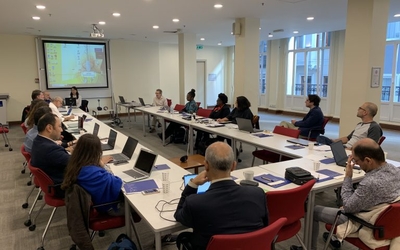 Participants sitting around conference tables in hollow rectangle with display screen in front.  