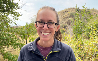 Dr. Joely DeSimone in blue fleece zippered jacket holding bird with trees and plants in background. 