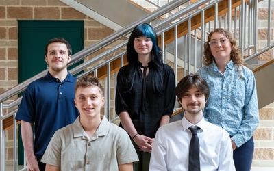 Two male interns seated with one male and two female interns standing behind them. Metal stairway and brick wall in the background. 