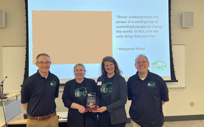 REACT's Michael Fiscus, Lesa Edwards, Jessica Mellon and Barry Hartung with award.  All are wearing REACT navy blue polos with green logos and are standing in front of slide screen with Margaret Meade quote in background. 
