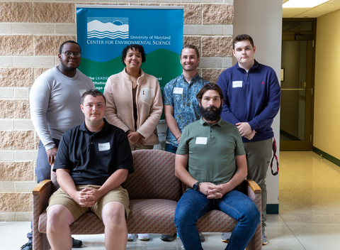 Six Appalachian Laboratory interns. Four standing behind two seated on pink sofa. Brick colored stone wall in background with UMCES logo. 