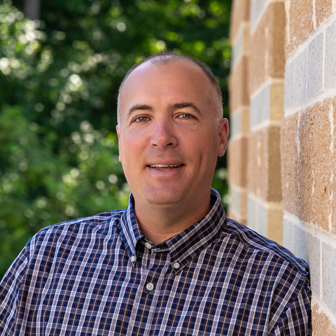 Headshot of Dave Nelson in blue and white checked shirt leaning against a red brick wall.  