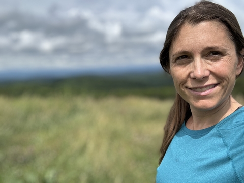 Selfie headshot of Ashley Dayer in blue shirt with blurred tall grasses in background.  