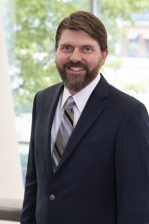 Andrew Elmore wearing a navy suit, white shirt and striped tie with a bright and sunny view of a tree behind him