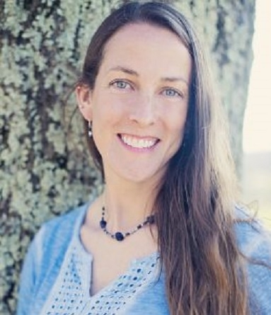 Katie Fallon headshot. Katie is wearing a blue and white top with matching necklace and is learning against a tree.  