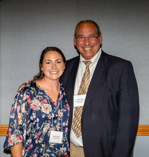 Marie Miller in blue flowered blouse standing beside Steve Resh in navy suit with tan striped tie with grey wall in background.