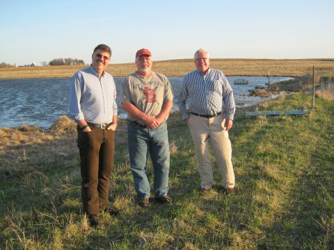 Three men pose on a green field with blue water in the background. 