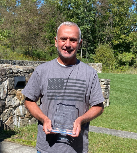 John Piasecki in grey t-shirt with US flag on it holding award plaque with green trees and rock wall in background. 