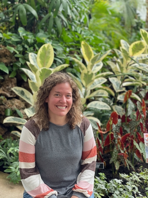 Annie Stark wearing a grey sweater with pink, white and grey striped sleeves with large green plants in background. 