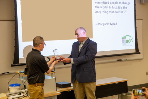 Mike Fiscus (REACT) accepting award from Dr. David Nelson (Appalachian Lab) in front of classroom with desks and screen in background. 
