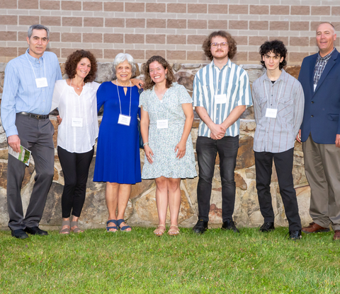Christopher Johnson, Marta Johnson Fiscus, Barbara Johnson, Annie Stark, Adron Fiscus, Julian Fiscus, and David Nelson standing and facing camera in front of grey stone and red brick wall. 