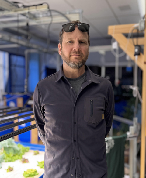 Headshot of Mick Burkett wearing a black button down shirt standing in an agriponics lab. 