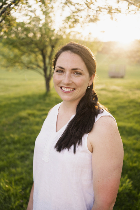Marie Miller dressed in white sleeveless top in a field of grass with tree and sun in background. 