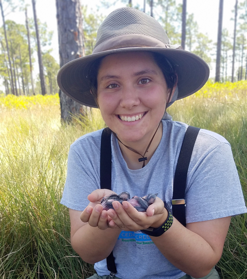 Megan Massa sitting in field of tall grass holding baby birds in both hands. 