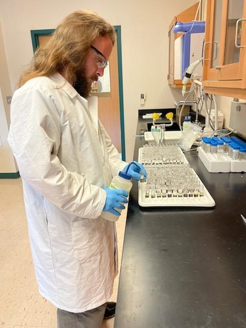 Richard Johnson wearing a white lab coat and holding plastic bottle while preparing samplesin testtubes in laboratory. 