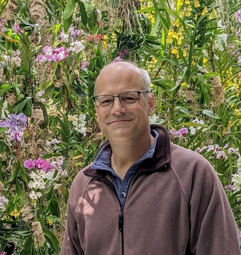 Dr. Tim Watkins in purple zipped sweatshirt standing in front of flowering trees. 