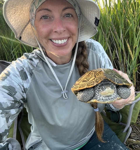 Photo of Jessica McGlinsey in a field of tall grasses wearing a wide-brimmed hat and holding a turtle