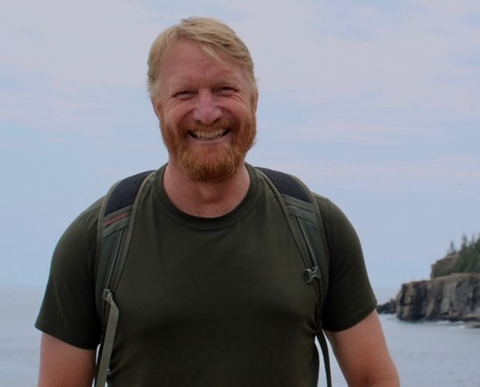 Brent Murry wearing dark t-shirt and backpack with coastline in background. 