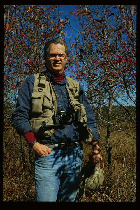 Dr. Richard A. Johnson outdoors surrounded by trees with binoculars and other birding equipment,