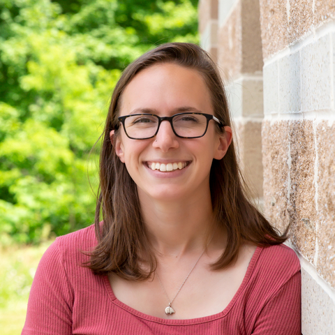 Headshot of Joely Desimone in red shirt leaning against red and white brick building with trees in background. 