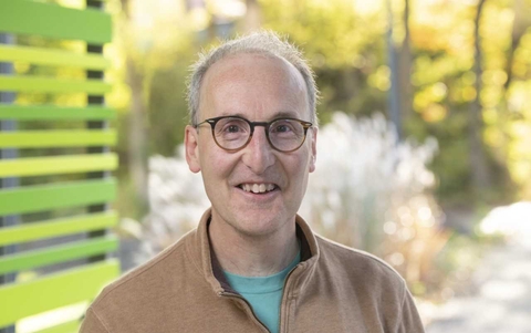James McClaren headshot, wearing a brown shirt over a turquoise t-shirt with blurred wooded scene in background 