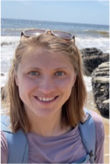Krista Kraskura in light purple shirt with backpack on the beach with waves in background.  