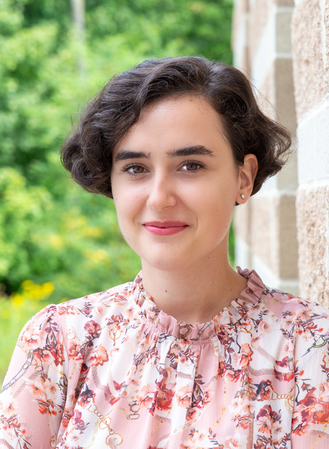 Haedshot of Mihaela Borota in pink floral blouse leaning against stone wall with blurred greenery in background. 