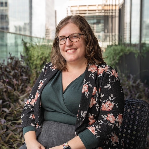 Woman with glasses and floral blazer sits on a balcony surrounded by plants.