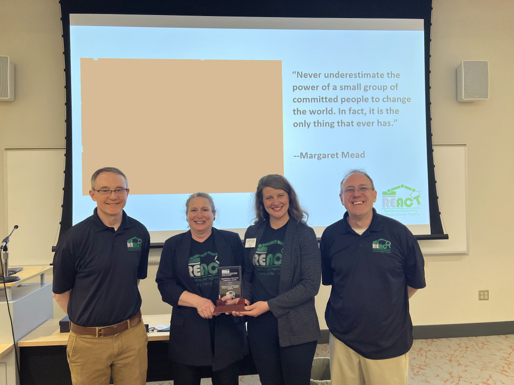 2023 Inspiration Award winner REACT: Michael Fiscus, Lesa Edwards, Jessica Mellon and Barry Hartung with award. All are wearing REACT navy blue polos with green logos and are standing in front of slide screen with Margaret Meade quote in background.