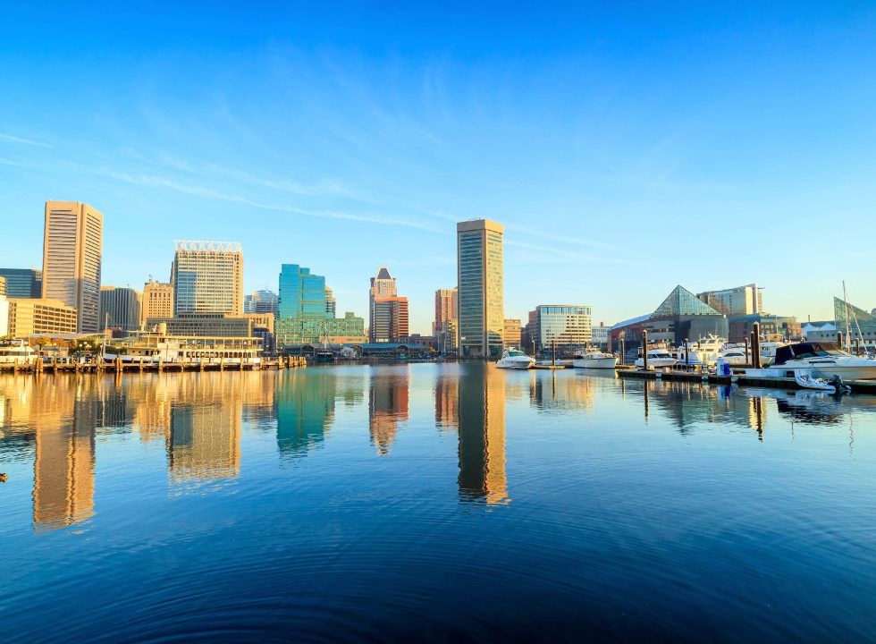 Baltimore Harbor skyline on a bright sunny day, with buildings reflecting in the still water of the harbor