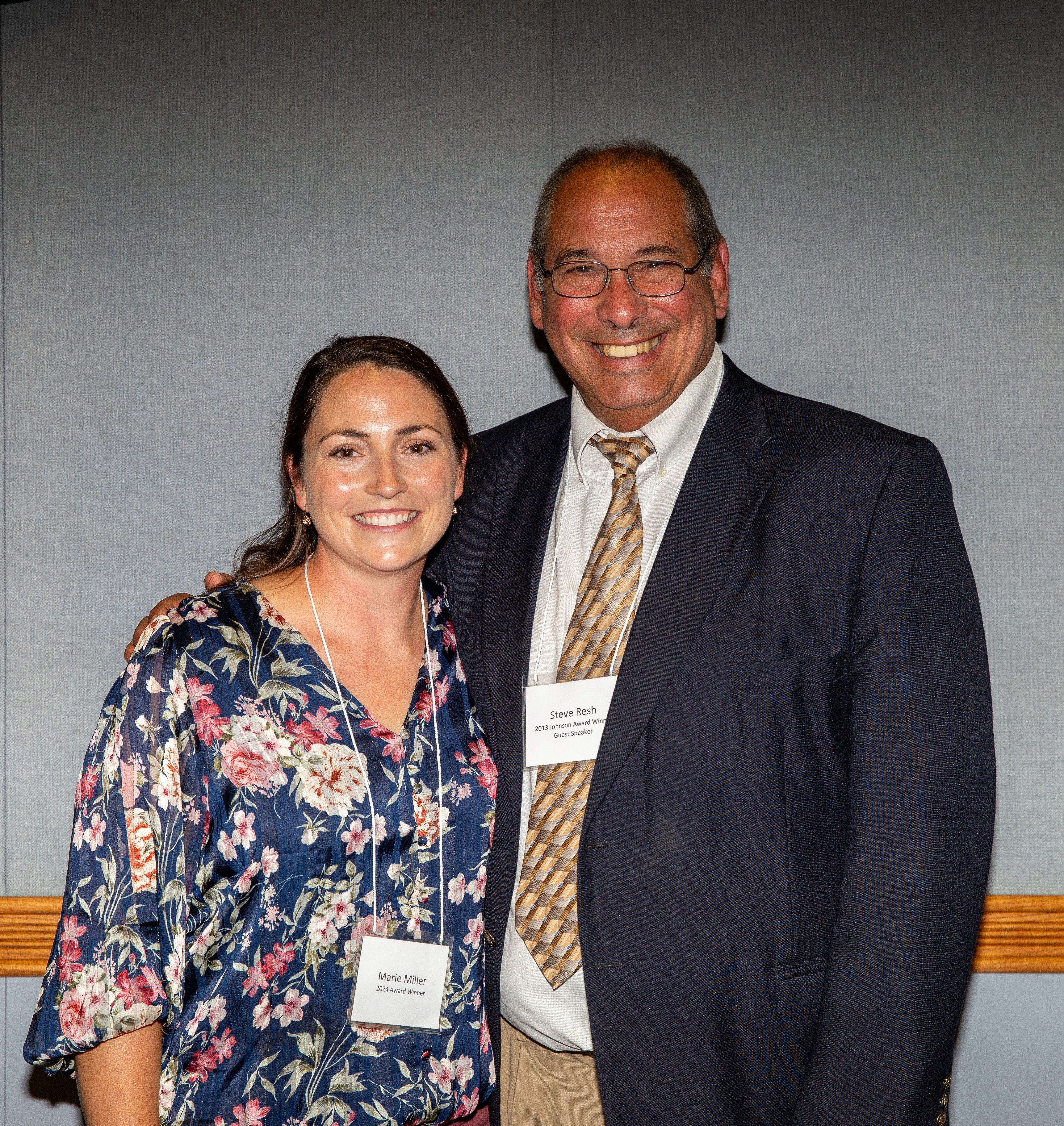 Marie Miller in blue flowered blouse standing beside Steve Resh in navy suit with tan striped tie with grey wall in background.