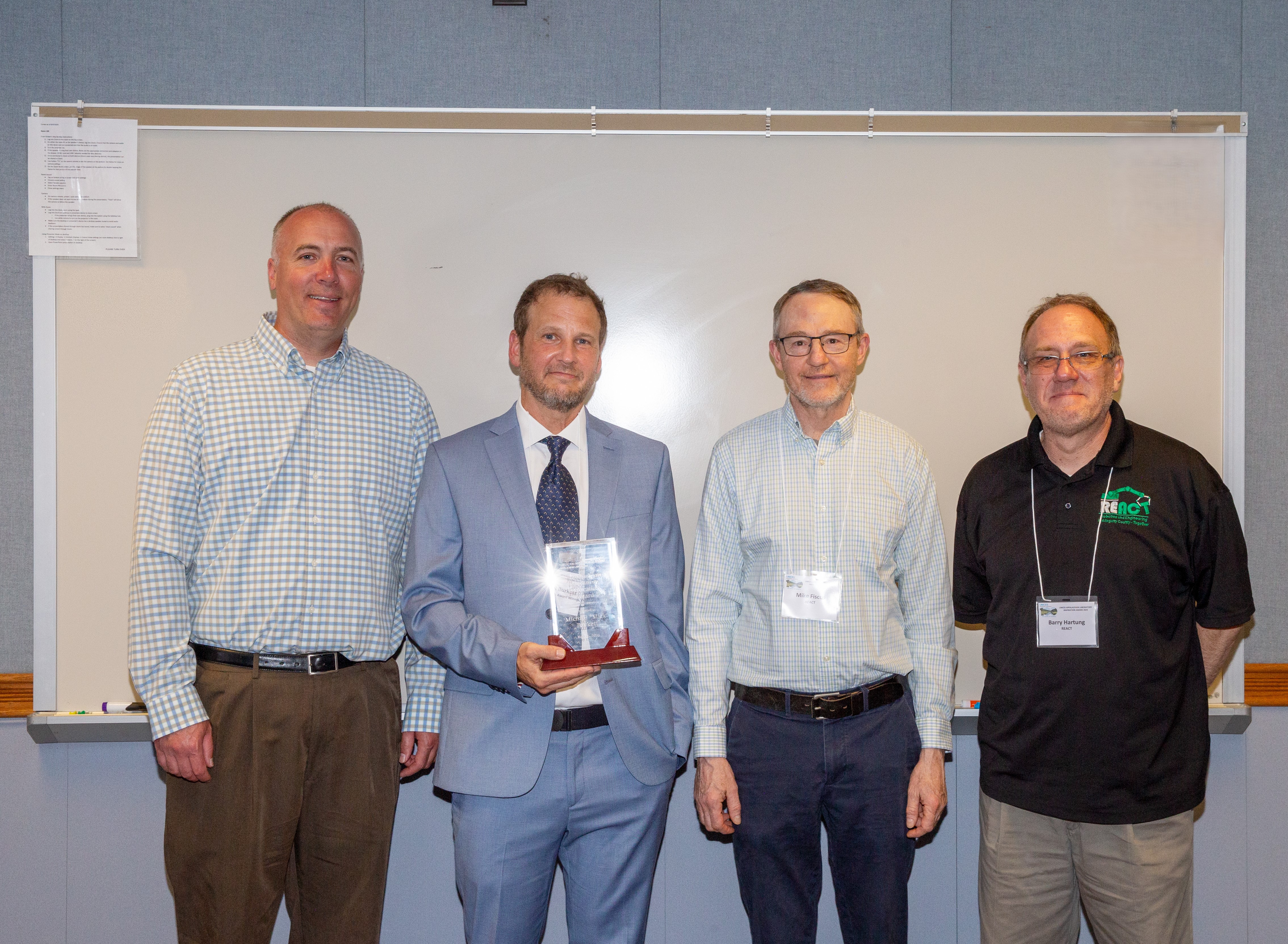 Dr. David Nelson, Mick Burkett, Mike Fiscus and Barry Hartung standing in front of white marker board, Mick is holding the award trophy.