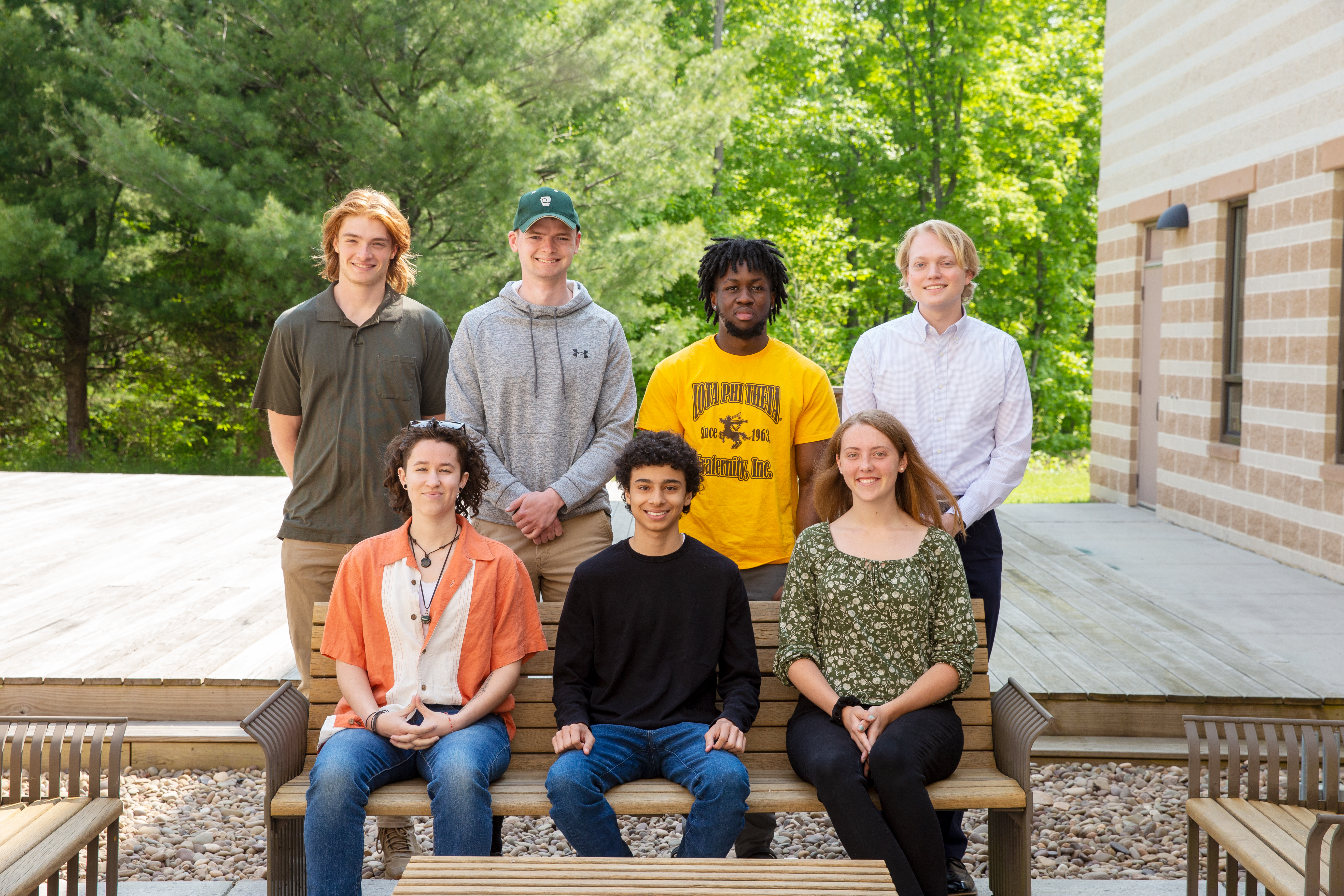 Group photo of 2023 cohort of interns outside with three interns seated on wooden bench and four standing in background with wooden deck and forest in background.