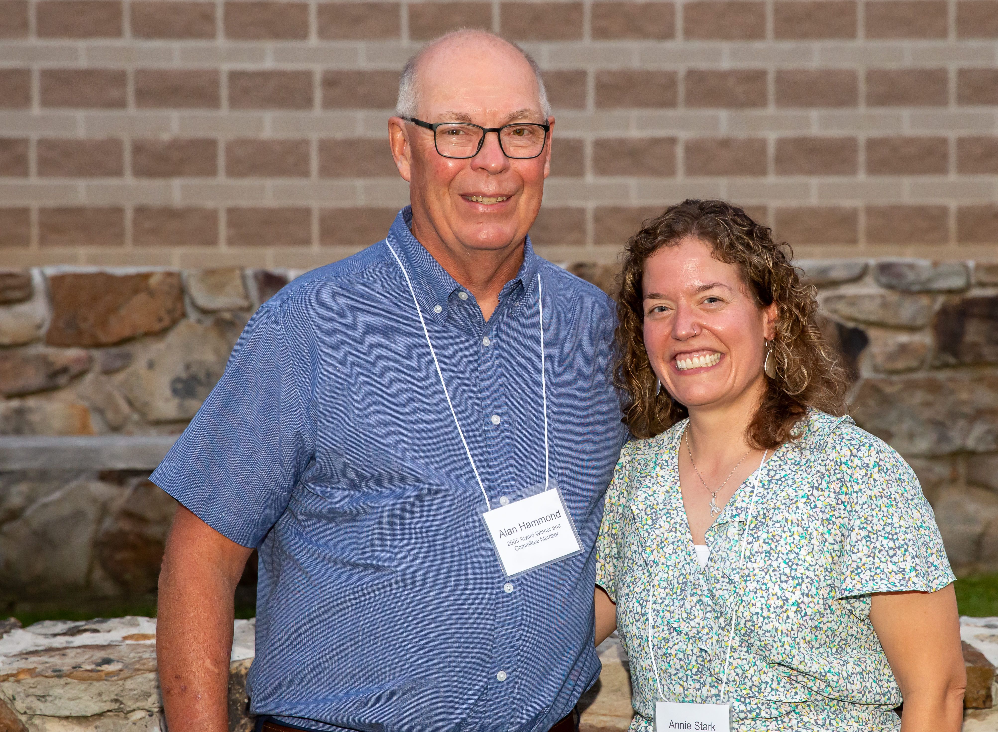 Alan Hammond in blue shirt standing next to Annie Stark in a light green flower patterned dress. Both are standing in front of a grey stone and red brick wall.