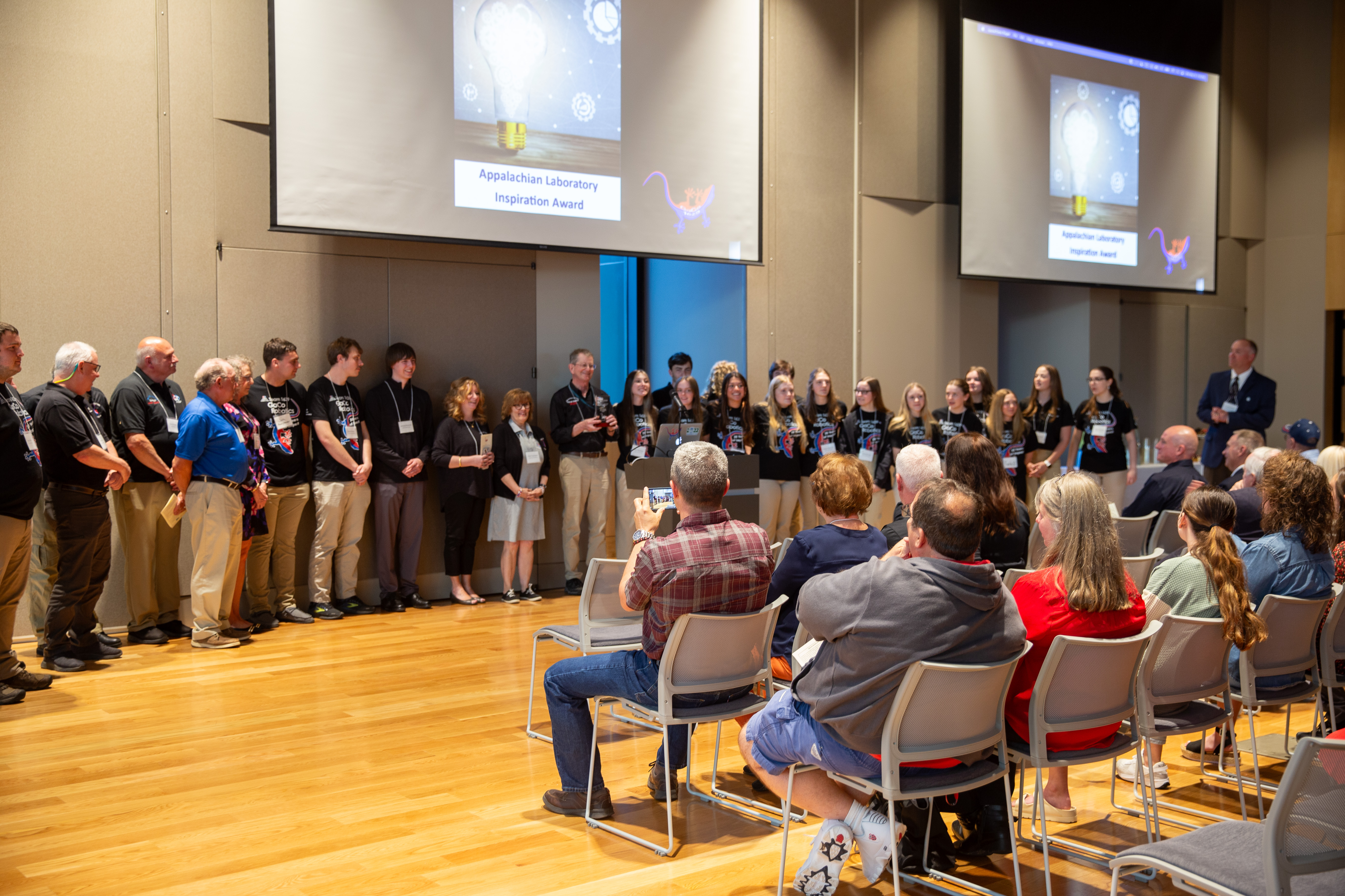 Inspiration Award event with numerous GaCo volunteers an students lined up with screens in background. Audience is seated in foreground with backs to camera.