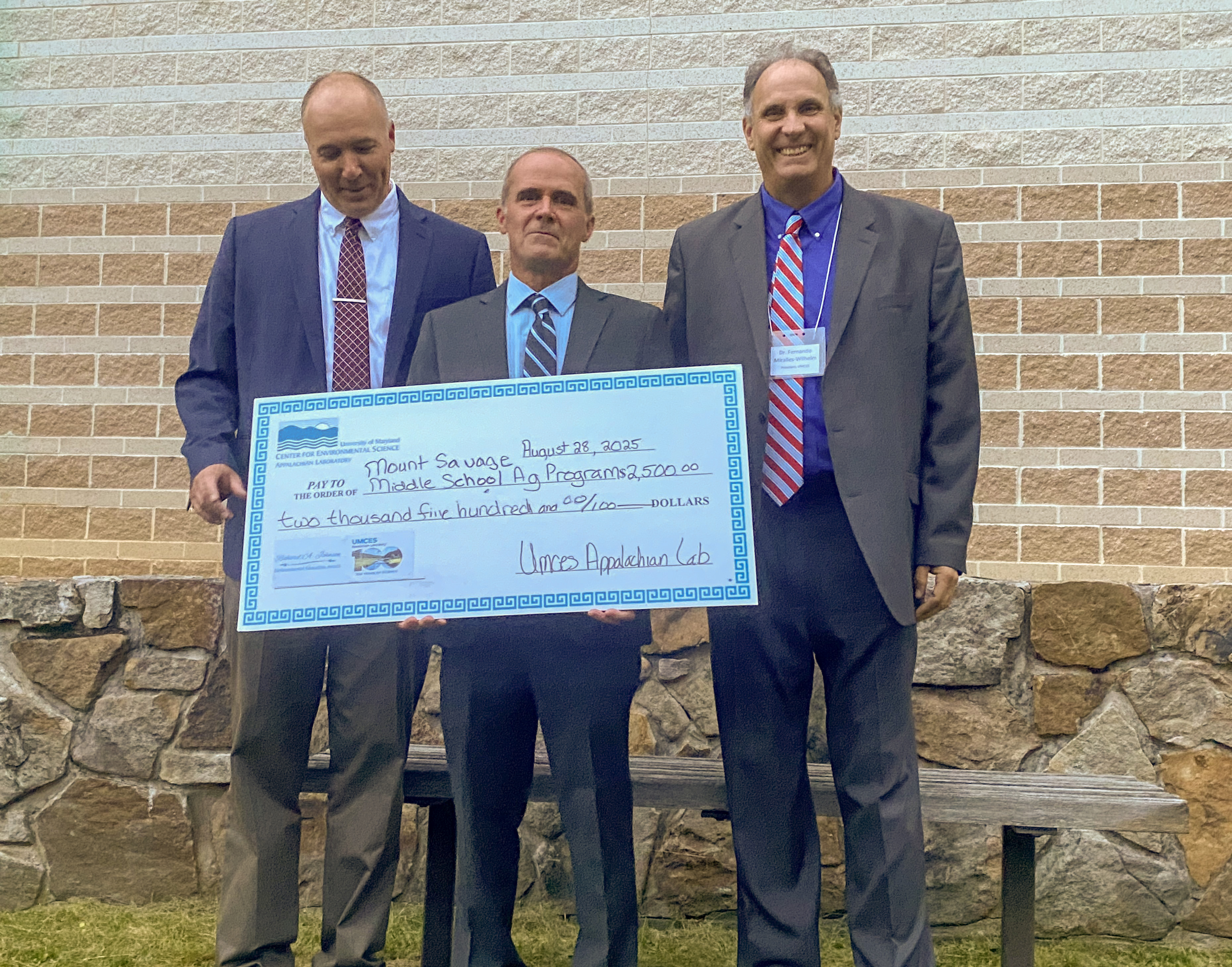 Dr. David Nelson, Matthew Kline, and Dr. Fernando Miralles-Wilhelm standing with prop check in front of stone wall with red brick wall in background.
